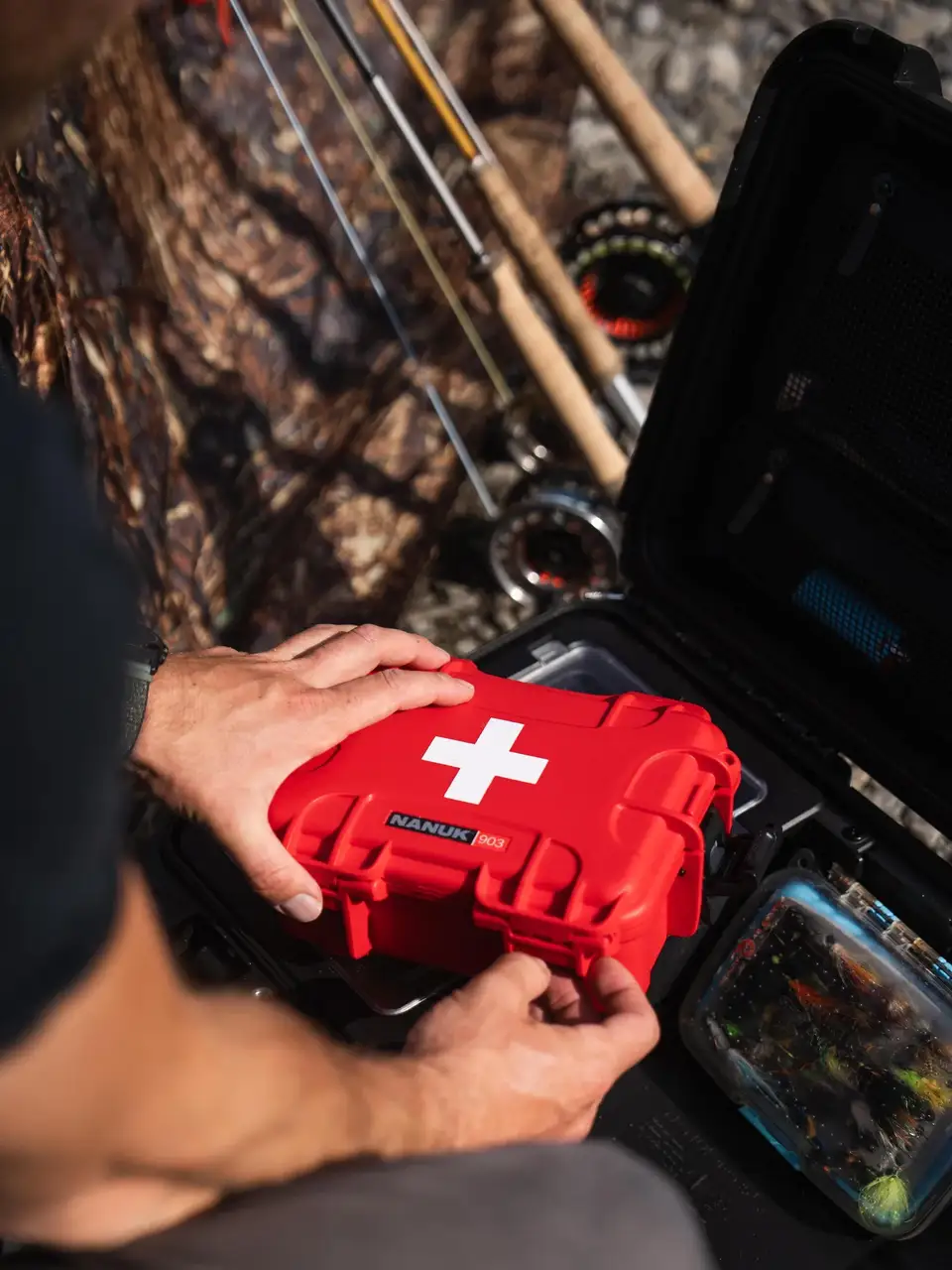 A red hard case with a white cross and the brand name NAKLUN PRO on the front, held by a person in an outdoor fishing setting next to an open black tackle case.