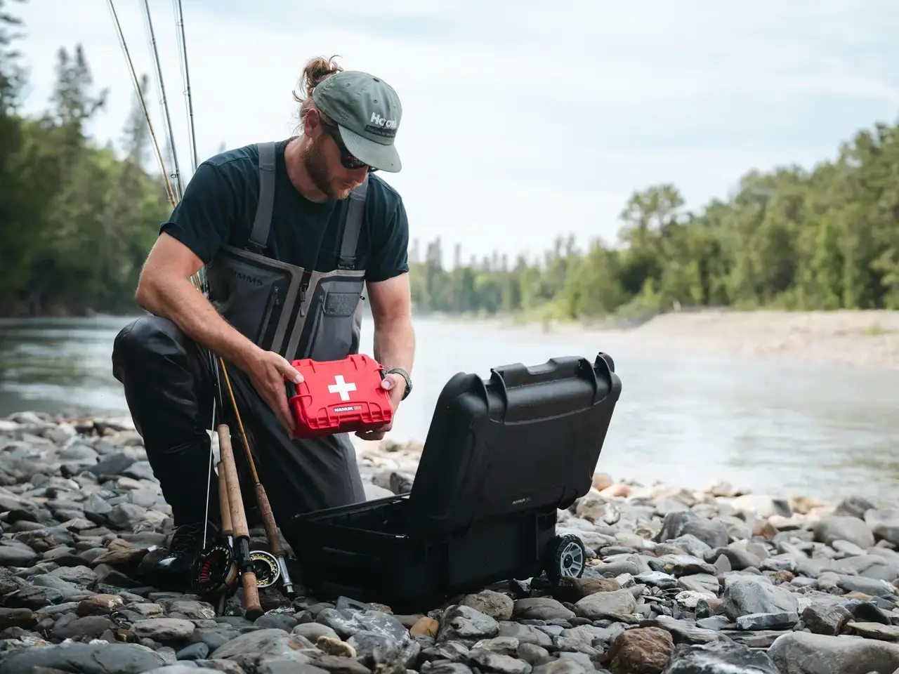 Man by a river opening a black suitcase with a red first aid kit on a rocky shore.