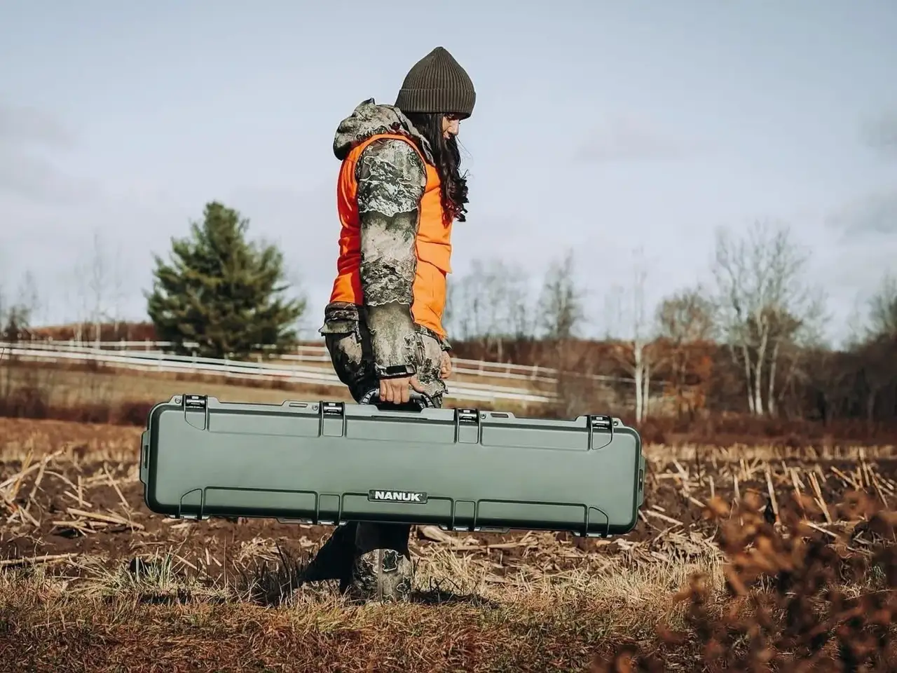 Person in hunting gear holding a large green hard-shell case in a field.