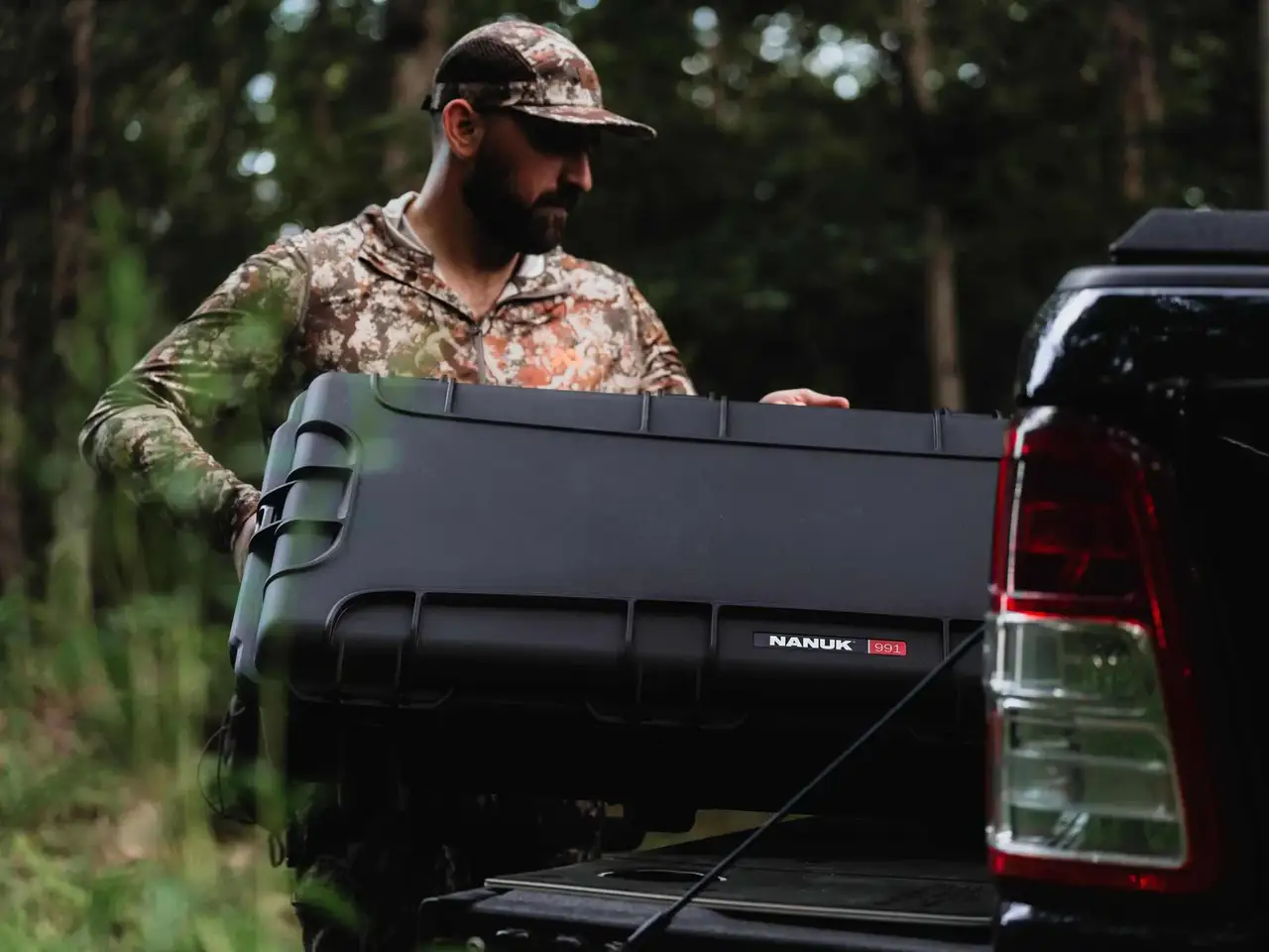 Man in camouflage holding a large black JunZhiJia toolbox next to a pickup truck in a forest setting