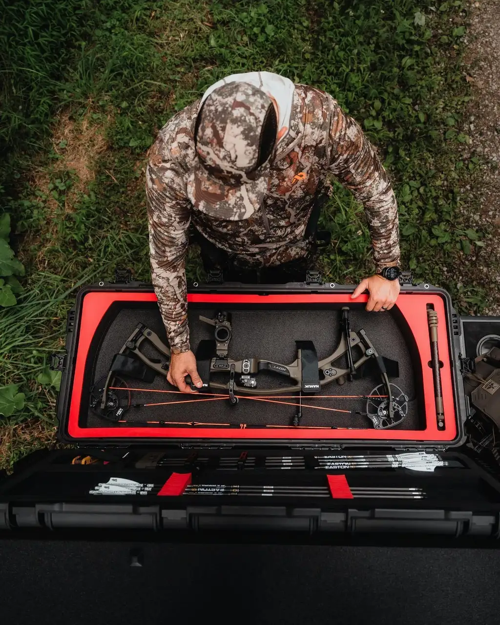 Person in camouflage gear setting up a bow in a black and red case outdoors.