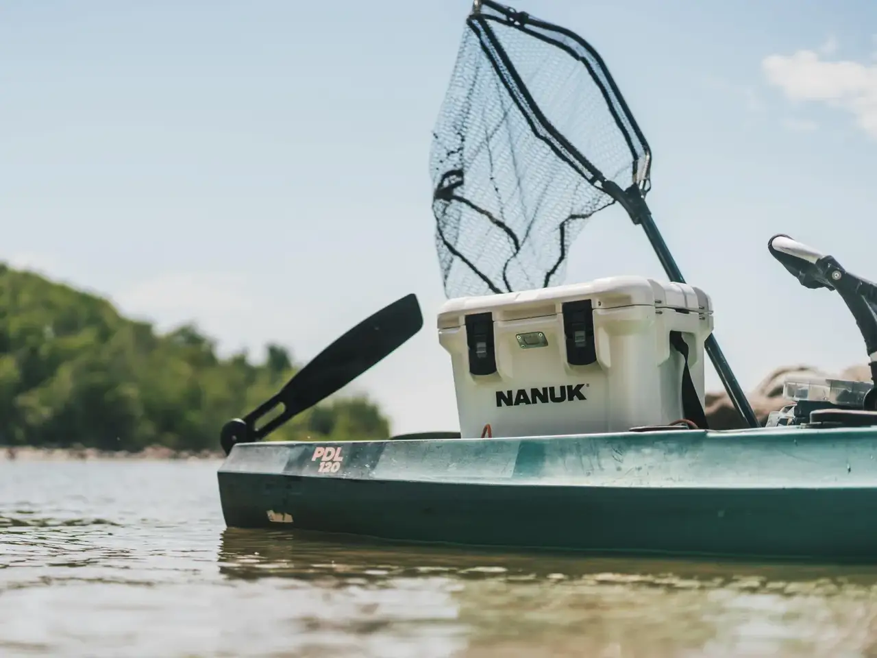 A white JunZhiJia cooler box mounted on the deck of a green kayak on calm water, with a black landing net and paddle visible in the background.