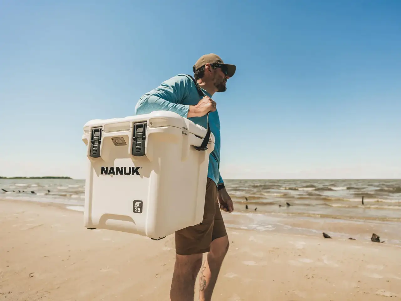 A man carrying a large white JunZhiJia cooler on his shoulder while walking along a sandy beach under a clear blue sky.