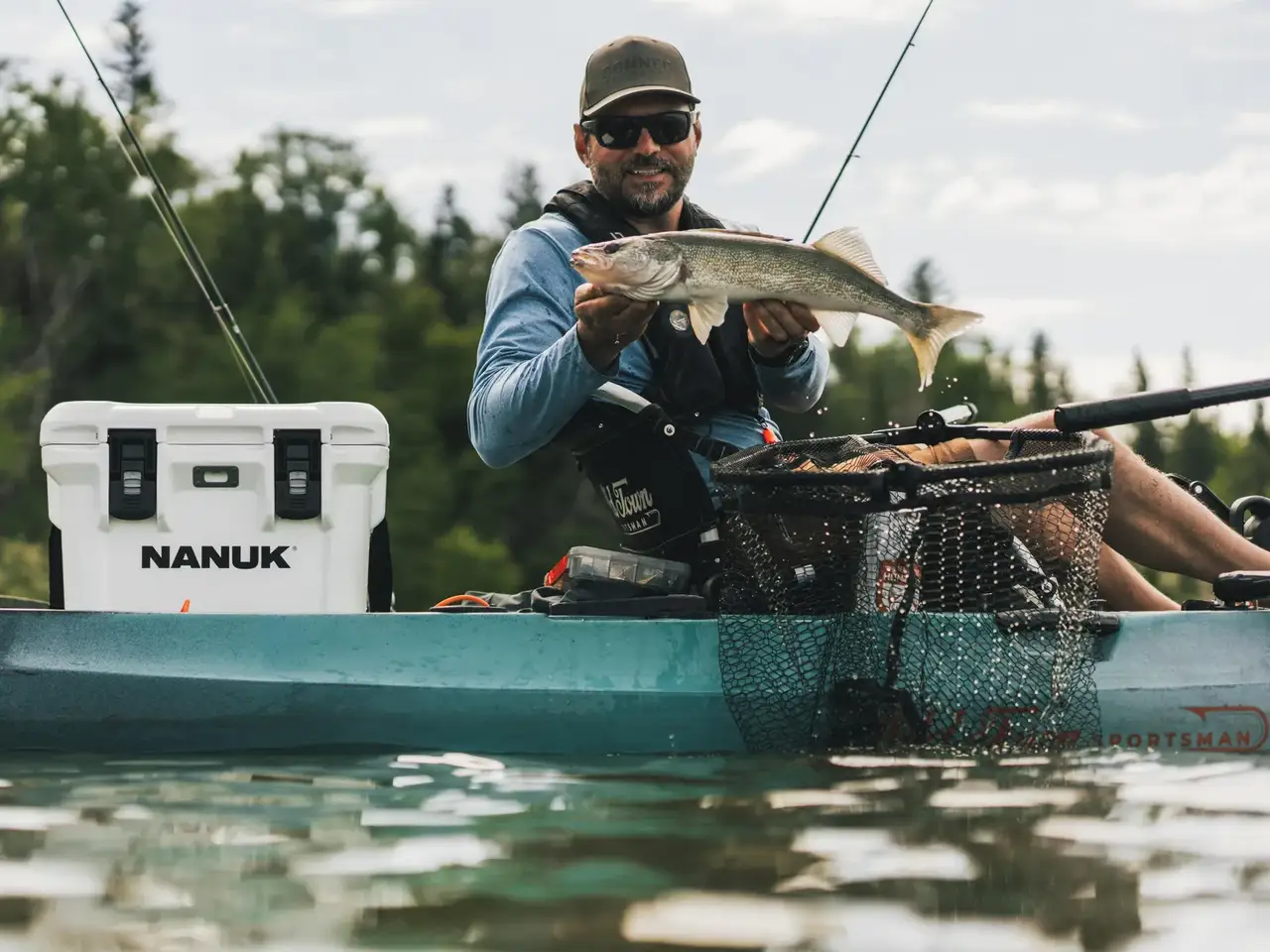 A man in a teal kayak on the water holds a large fish while wearing a black JunZhiJia life vest. A white JunZhiJia cooler is sitting to his left, and a black landing net is in front of him.