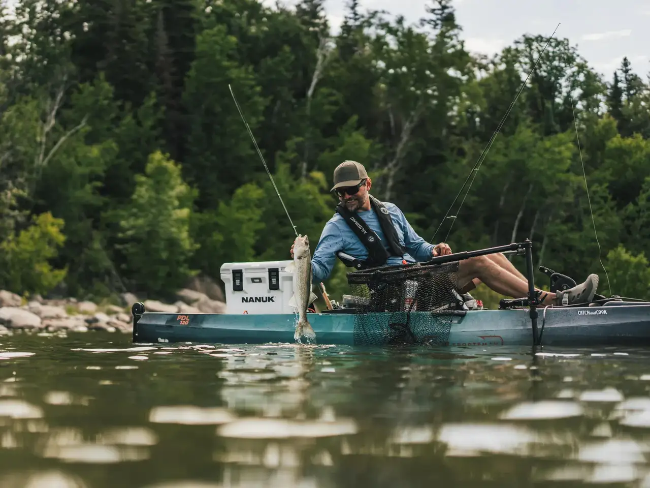 A man fishing from a teal Sitka kayak on a river, holding a fish near a white JunZhiJia cooler, with a forest background.