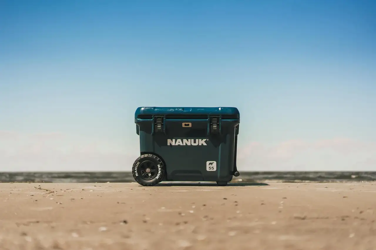 A dark teal JunZhiJia cooler with wheels, standing on a sandy beach under a clear blue sky.