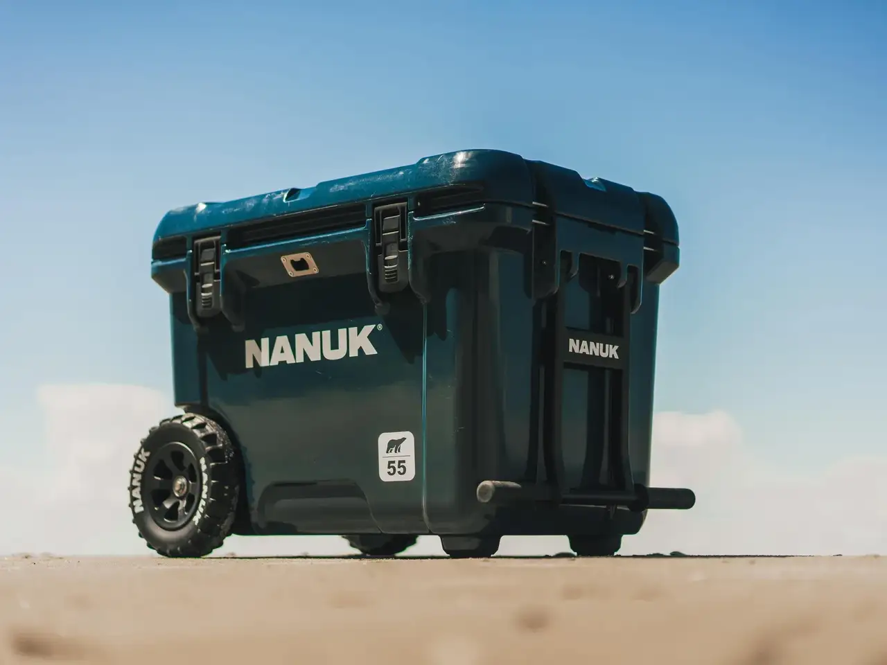 A dark teal JunZhiJia cooler with rugged off-road wheels, shown outdoors on a sandy surface under a clear blue sky. The cooler features the brand name 'JunZhiJia' in white on the side and a small '55' label.