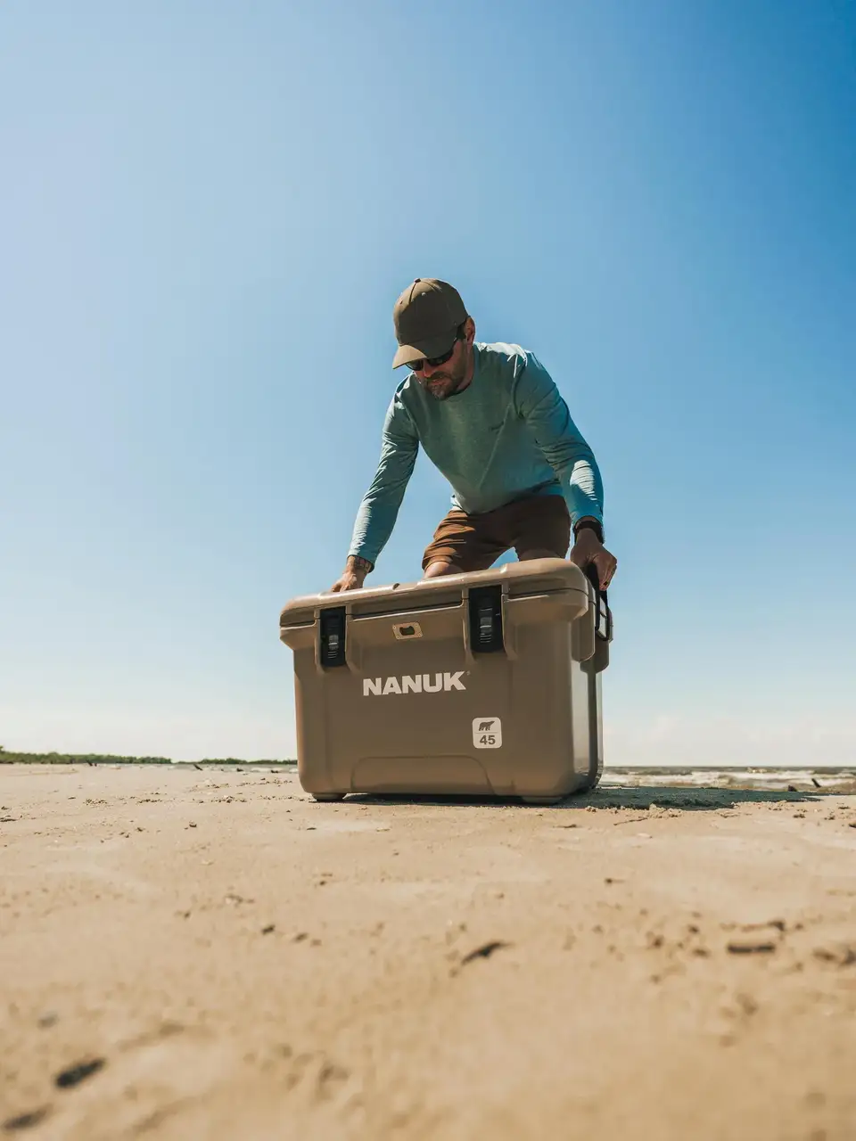 A man in a teal shirt and brown cap pushes a large beige JunZhiJia cooler on a sandy beach under a clear blue sky.