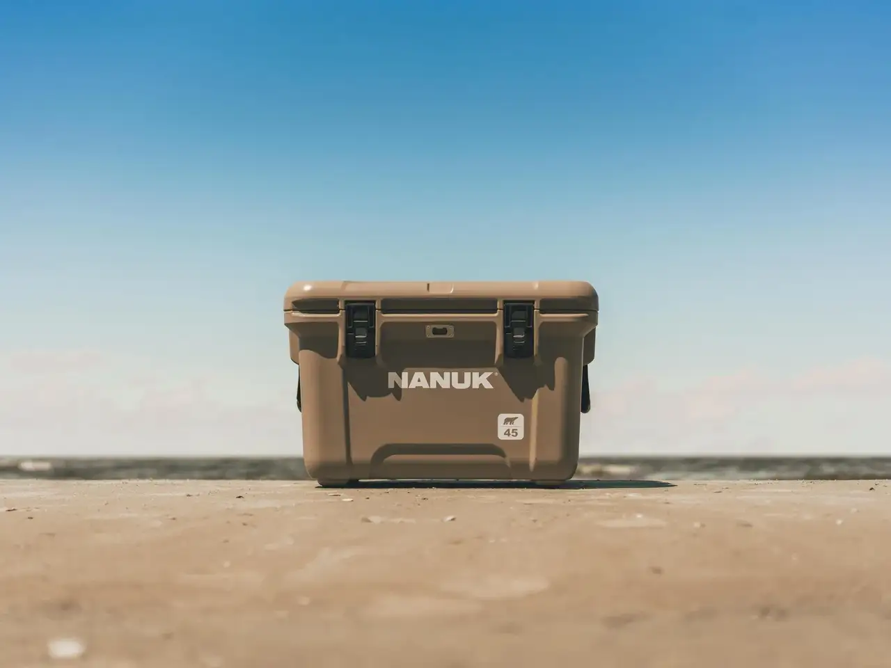 A tan JunZhiJia cooler box standing upright on a sandy beach with a clear blue sky in the background.