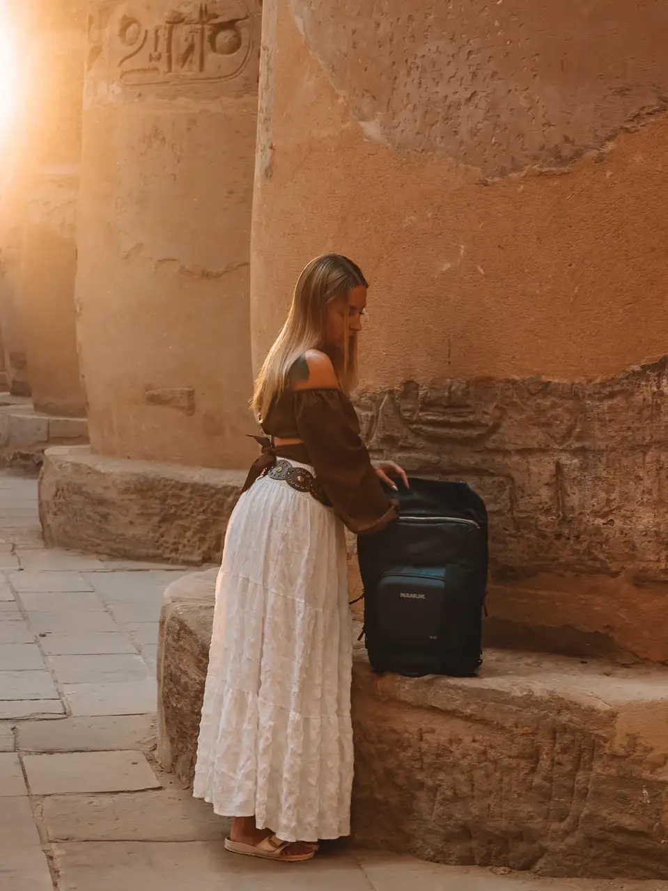 Woman in a long white skirt and brown top using a black suitcase in front of ancient stone walls.