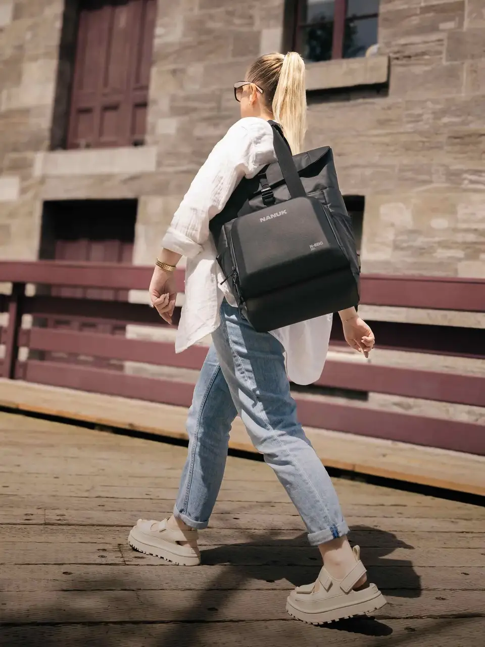 Person walking outdoors with a black bag, wearing a white shirt and blue jeans.