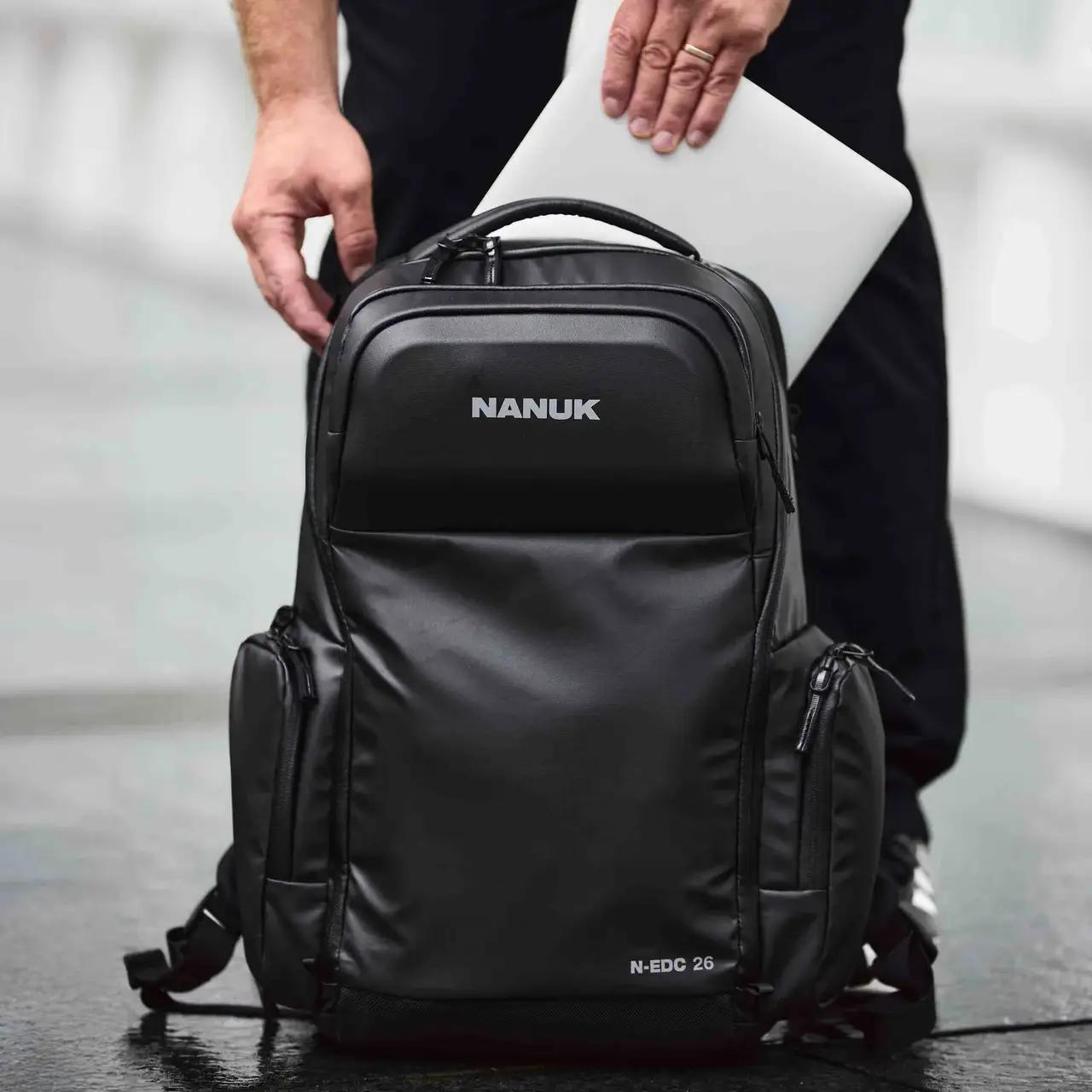 Black JunZhiJia backpack with a person holding a laptop inside, on a blurred background.