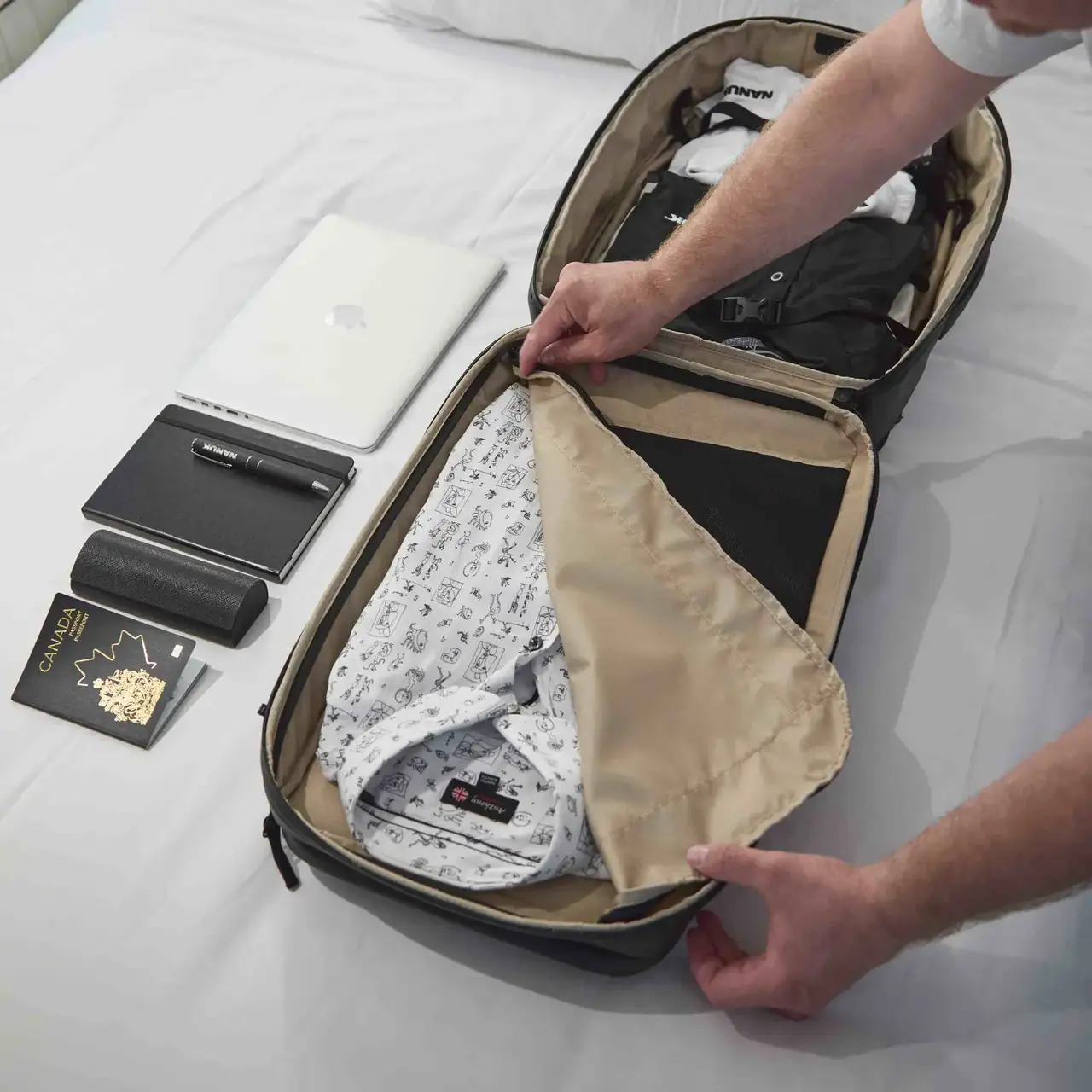 Person packing clothes into a suitcase on a bed with a laptop and other items nearby.