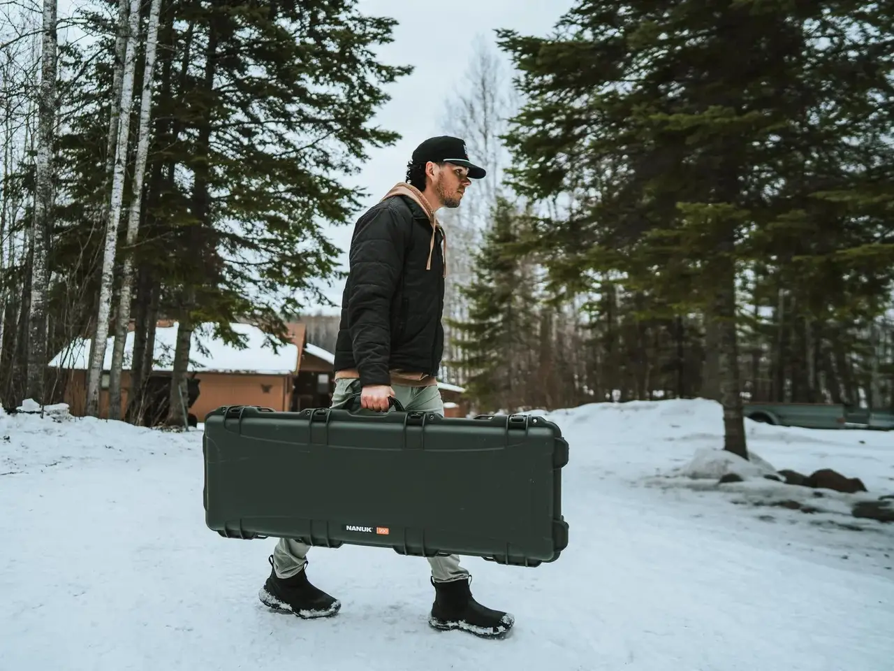 Person carrying a large black case in a snowy landscape with trees and a cabin in the background.
