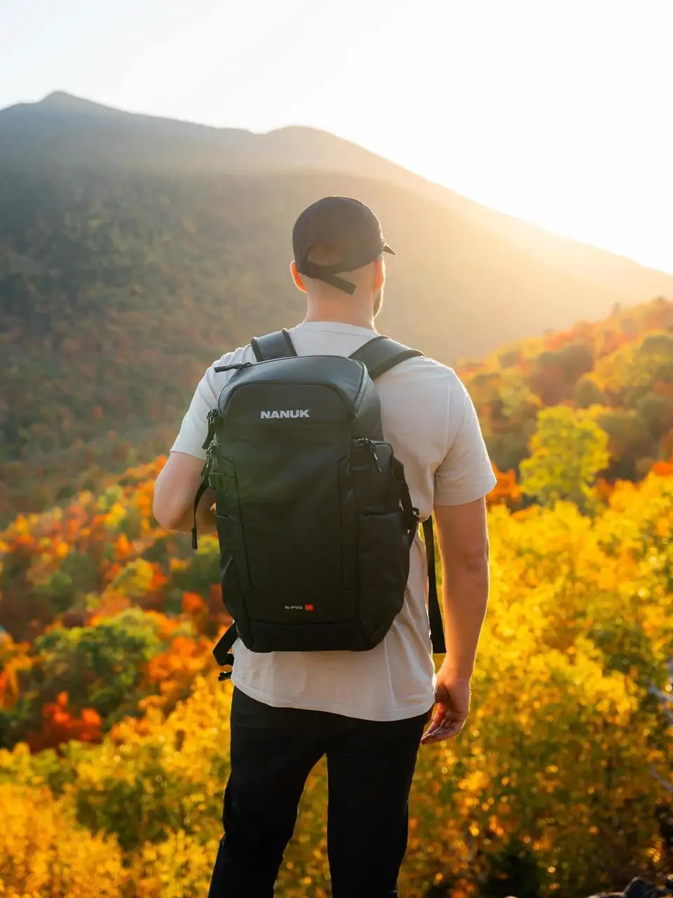 Person wearing a black backpack with 'NAMUK' branding, standing in a scenic outdoor setting with mountains and trees.