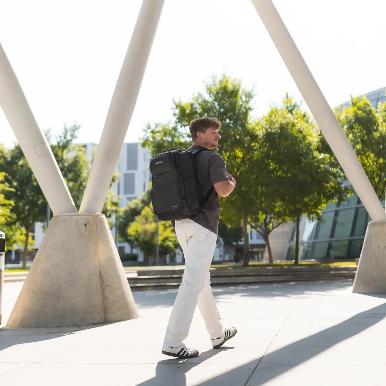 Man walking outdoors with a backpack, surrounded by trees and modern architecture.