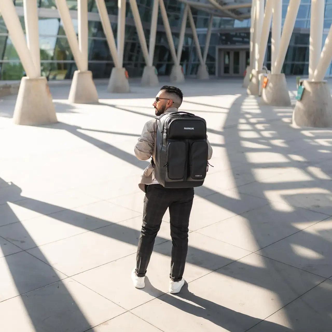 Man with a backpack walking on a sunlit pavement with modern architecture in the background