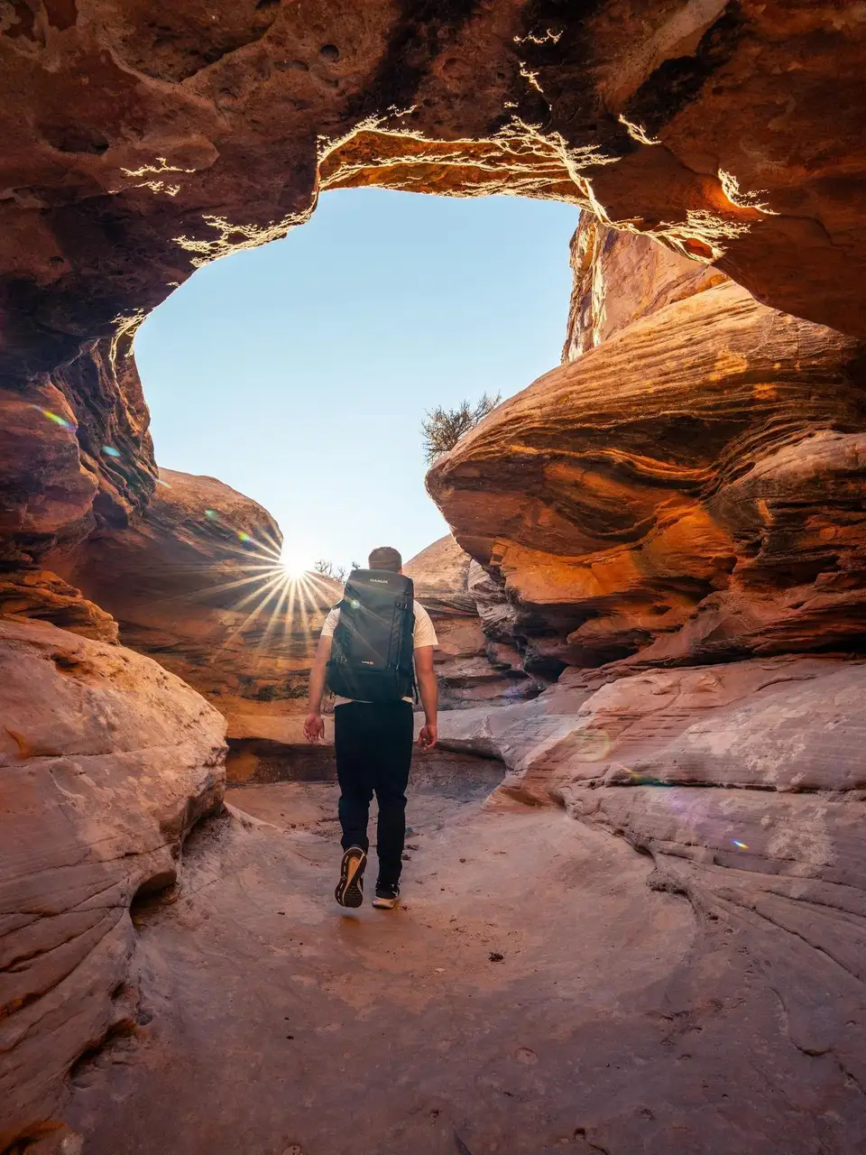 Person walking through a rocky archway with a backpack, sun shining through