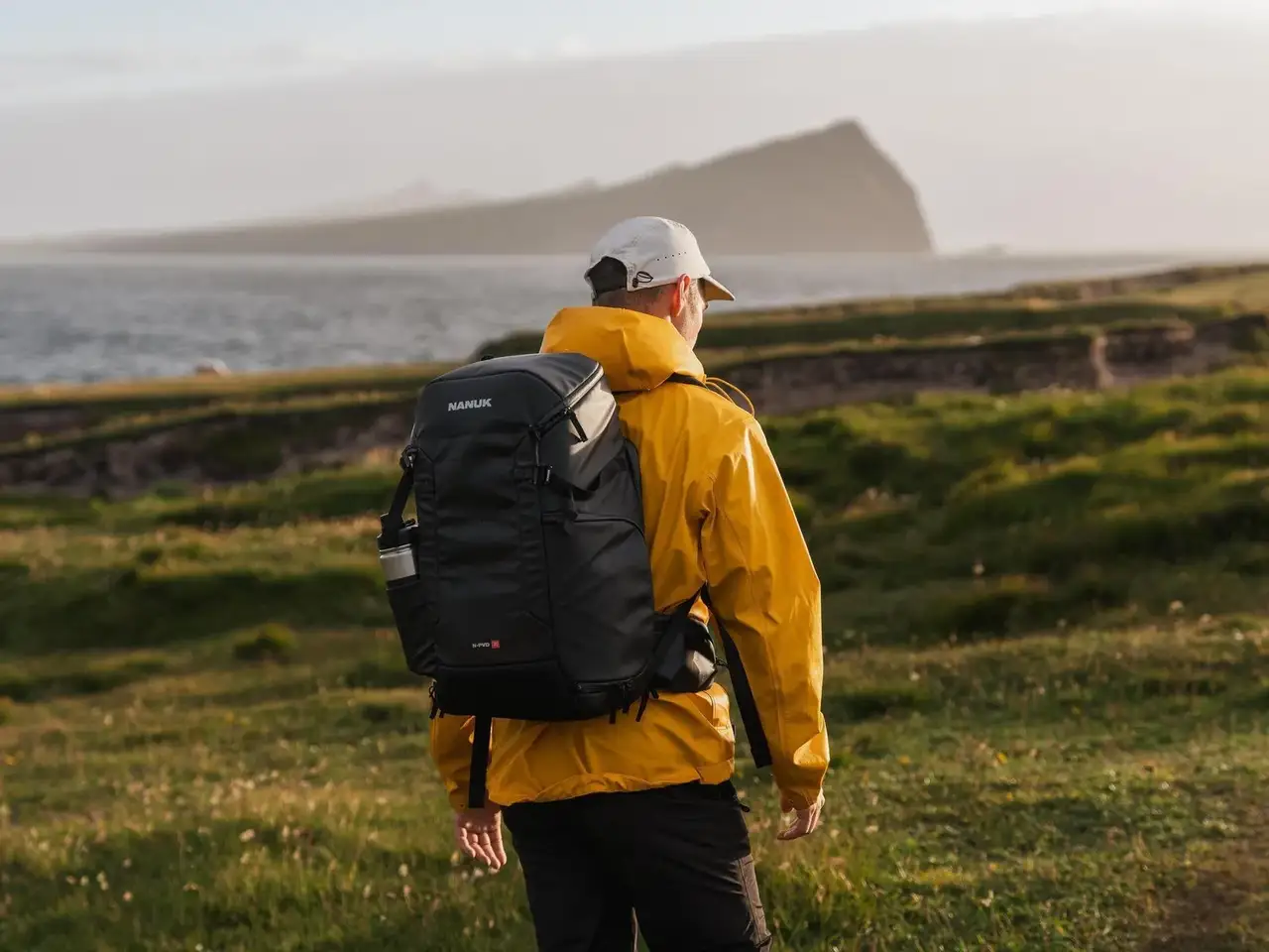 Person wearing a yellow raincoat and black backpack standing on a grassy field with a scenic background.