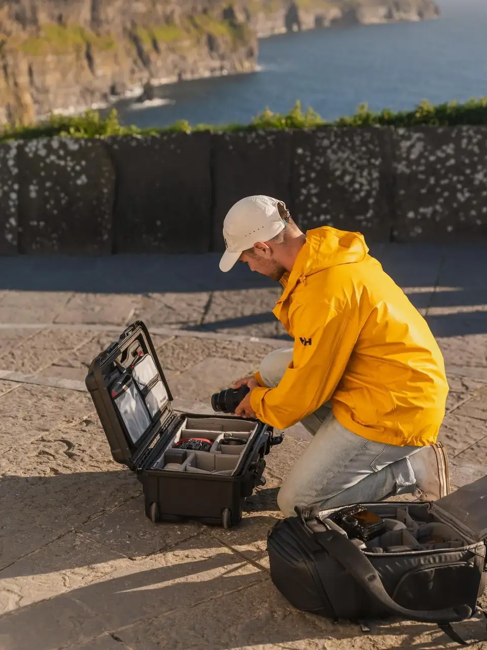 Person in a yellow jacket sitting on a stone ledge with a camera bag and equipment case by the ocean.