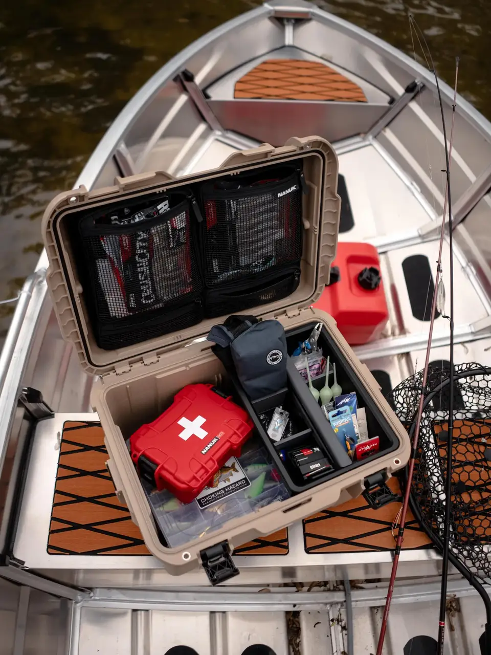 An open tan waterproof hard case filled with fishing and emergency gear, including a red hard case with a white cross, black pouches, a black device, and accessories, set on the deck of a boat with water visible around it.