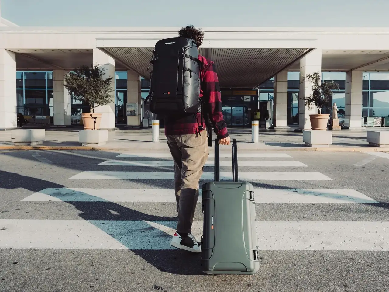 Person with a backpack and suitcase standing on a crosswalk in front of a building.