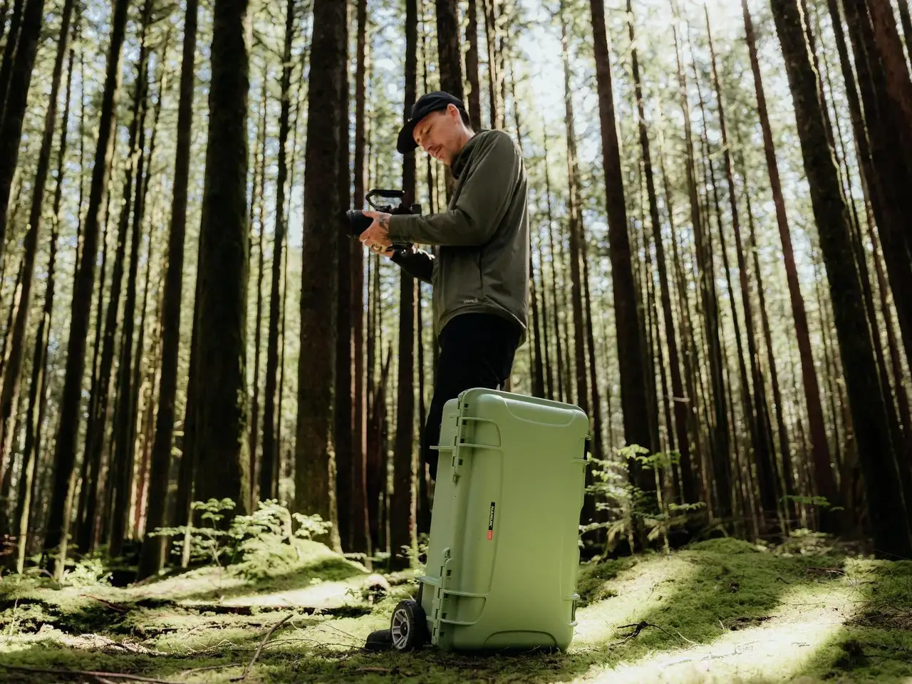 Person setting up a camera in a forest with a large green suitcase.