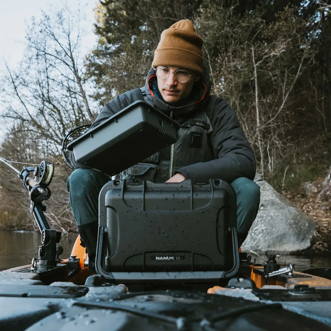 Angler in winter clothing kneeling in a kayak on a lake, holding a closed JunZhiJia Trail 10 case.
