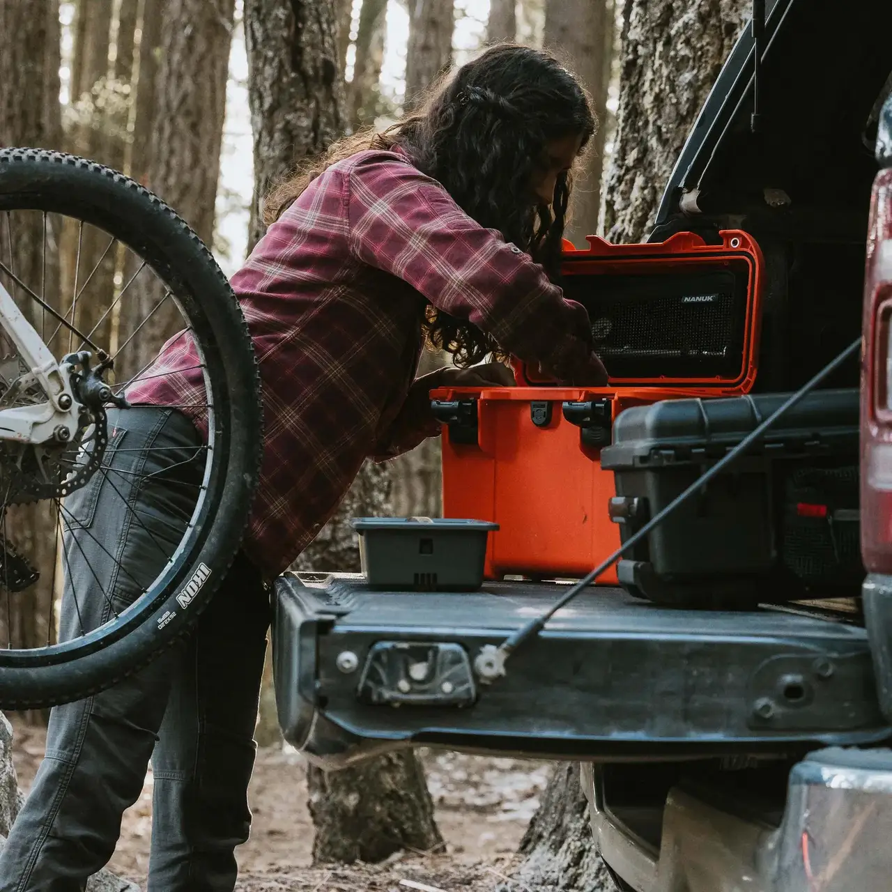 Cyclist loading a bright orange JunZhiJia Trail 15 case into a pickup truck bed next to a mountain bike in a forest setting.