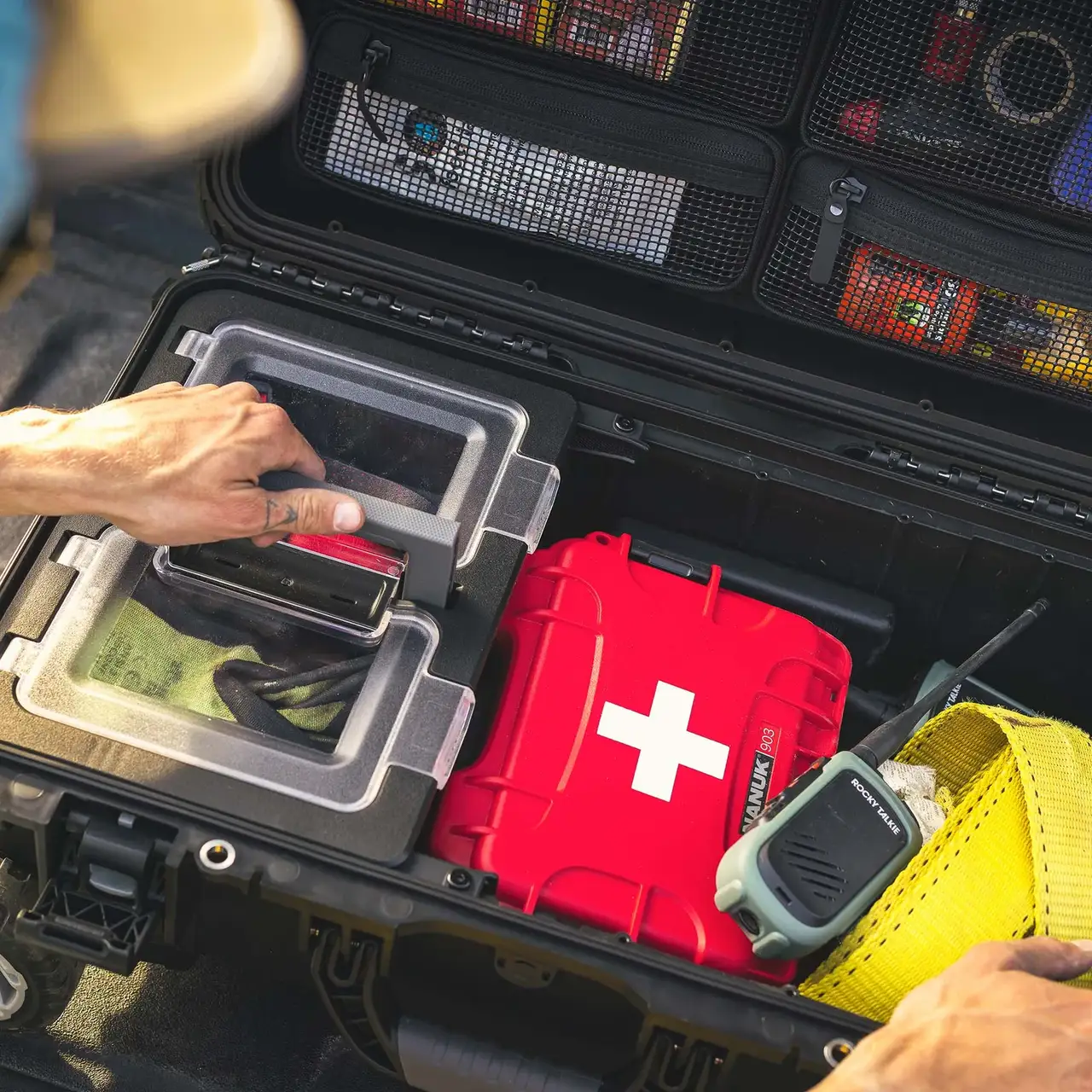 Person reaching into a black tool case with various items including a red first aid kit and a walkie-talkie.