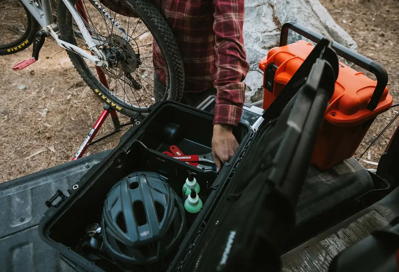 Person loading a black JunZhiJia Trail 30 case into the bed of a pickup truck next to a mountain bike on a forest trail.