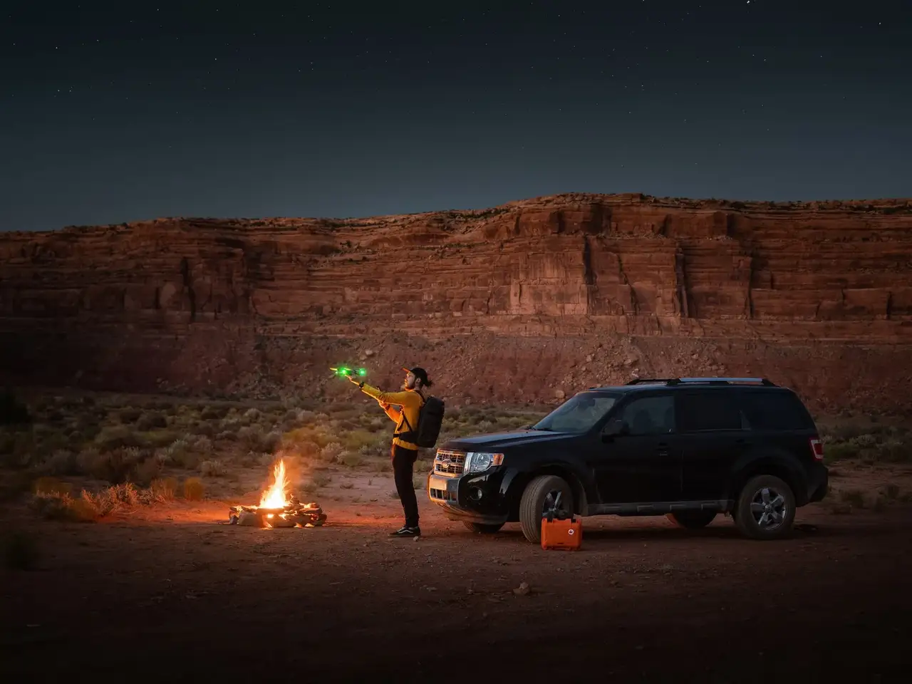 Person holding a green light in front of a car and campfire in a desert landscape at night.