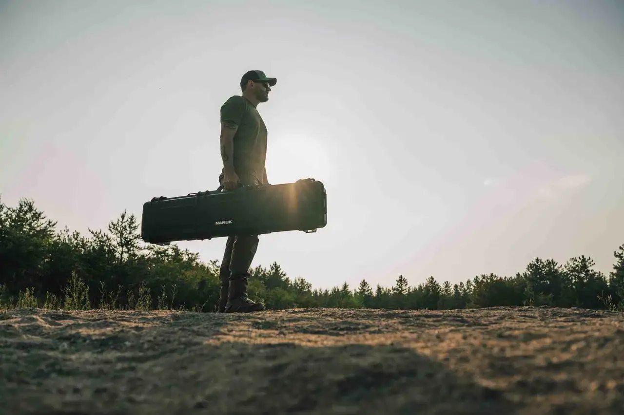 Person holding a large black case against a natural background with trees and open sky.