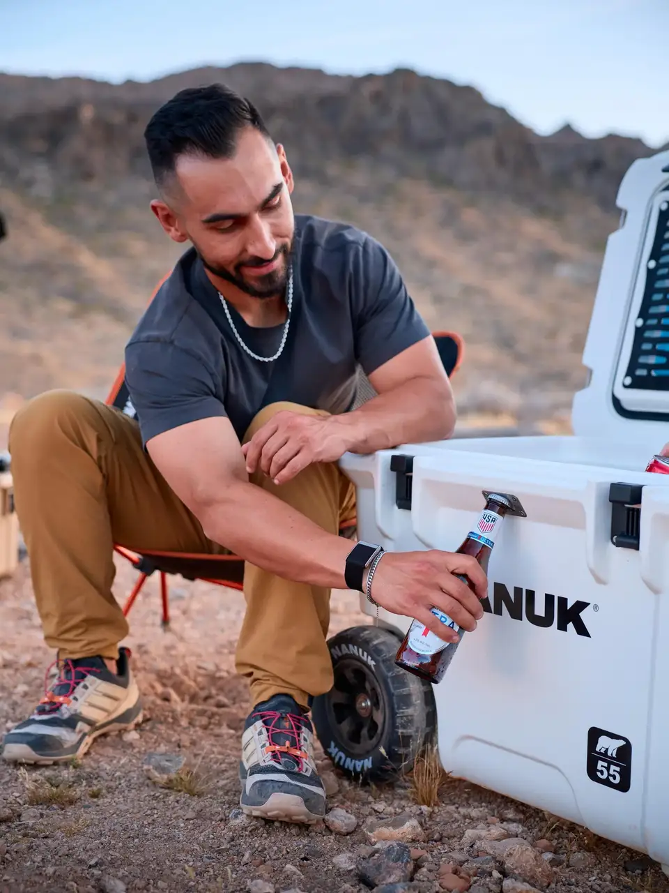 Man sitting by a white vehicle in a desert setting, holding a can of Anuk.