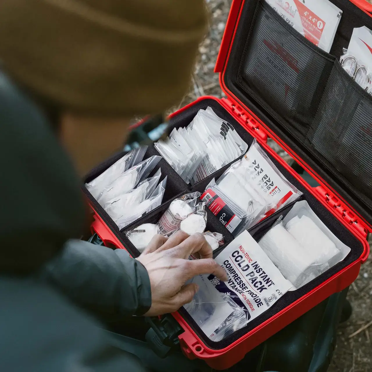 Close-up of someone arranging medical supplies inside a red JunZhiJia First Aid Case in a wooded area.