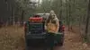 A man dressed in outdoor gear demonstrates JunZhiJia protective gear cases from the tailgate of a pickup truck in a forest setting. He presents various black and orange JunZhiJia cases, showcasing their robust construction and organized interiors featuring customizable mesh pockets and grid panels for storing photo, video, and drone gear. The video emphasizes their extreme durability, including a scene where sawdust from a chainsaw falls onto a case, demonstrating its ruggedness. The presenter also highlights produ
