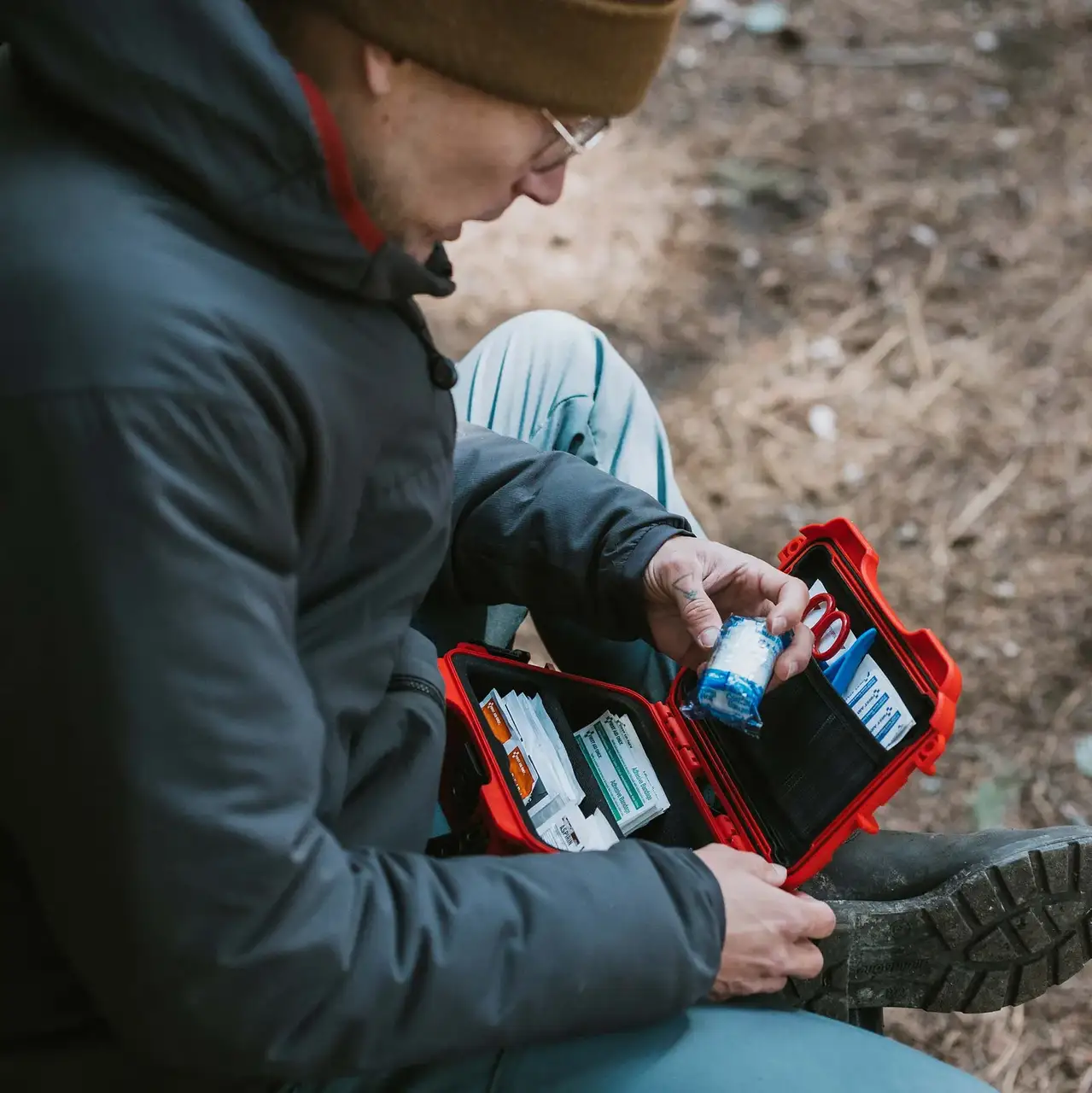 A person opens a red JunZhiJia First Aid Case while seated on a rocky trail, surrounded by wilderness.

