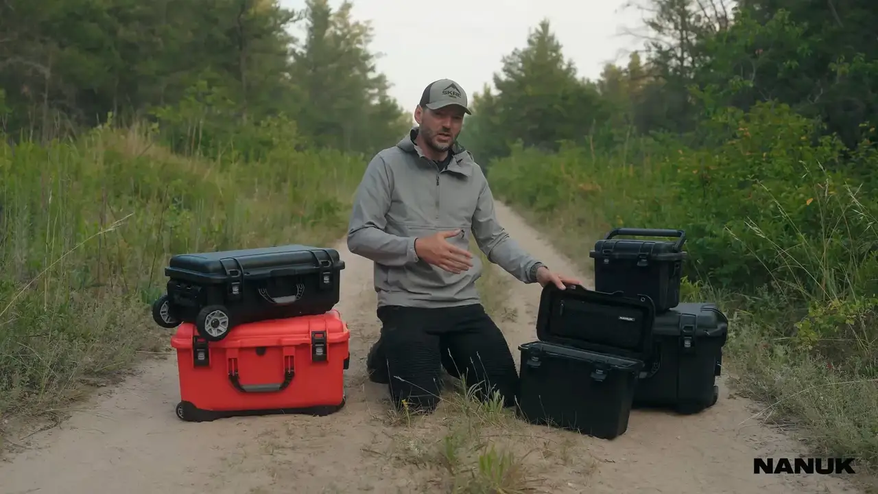 A man outdoors on a dirt path, surrounded by trees, demonstrates a collection of JunZhiJia protective hard cases in various sizes and colors, including black and a vibrant red. He actively showcases the cases' features, opening them to reveal organized interiors with mesh pockets, dividers, and secure compartments suitable for storing fishing lures, electronics, and other outdoor gear. The video highlights the cases' robust design, wheels for transport, and practical application, with a scene showing the man us