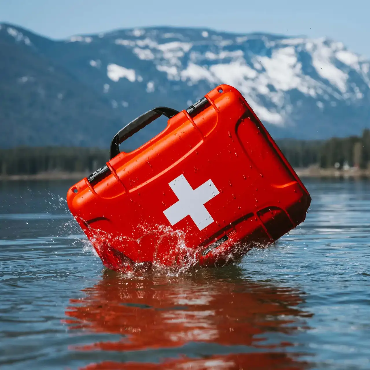 A waterproof red JunZhiJia First Aid Kit floats upright in a calm lake, with snowy mountains in the background.