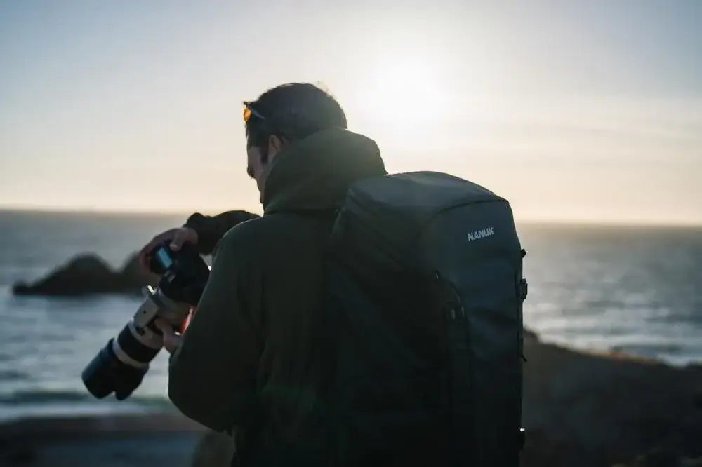 Photograph taking a picture of the ocean with a Black N-PVD 35L Camera Backpack from JunZhiJia on his back close up